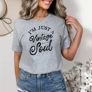 Woman wearing a gray t-shirt with 'I'm Just a Vintage Soul' text, standing against a neutral background with flowers.