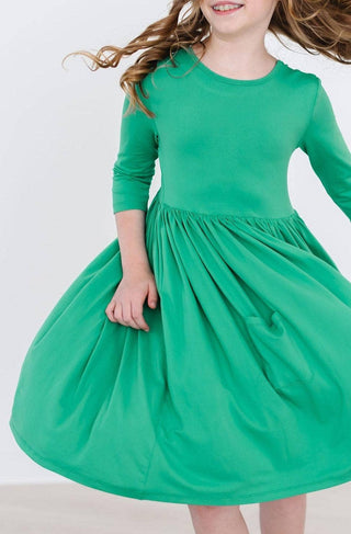 Toddler girl wearing a Kelly green 3/4 sleeve twirl dress with pockets from JSQ Mercantile – a women’s boutique and gift shop in La Grange, IL.